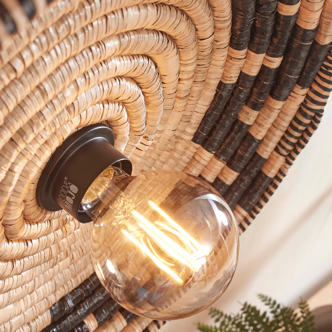 Ein Bild aus dem Taiga Store von taiga.green. Eine Nahaufnahme der Deckenlampe Andros (Durchm.45x8cm), einer geflochtenen, runden Deckenleuchte mit schwarzen und naturfarbenen Wirbeln, leuchtet warm. In der rechten unteren Ecke ist teilweise eine kleine Grünpflanze zu sehen. Jetzt bestellen im Eco Shop von www.taiga.green. Hier nachhaltig einkaufen mit dem Taiga Eco Rating