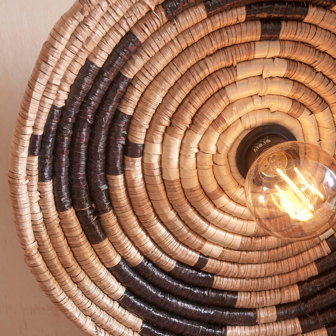 Ein Bild aus dem Taiga Store von taiga.green. Nahaufnahme der Deckenlampe Andros Diam.45x8cm, einer runden, geflochtenen Deckenleuchte im Korbstil mit konzentrischen Kreisen in natürlichen und dunklen Tönen und einer freiliegenden Glühbirne in ihrer Mitte. Jetzt bestellen im Eco Shop von www.taiga.green. Hier nachhaltig einkaufen mit dem Taiga Eco Rating