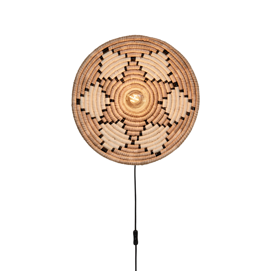 Ein Bild aus dem Taiga Store von taiga.green. Die Wandlampe Andros Diam.45x8cm hat einen runden, geflochtenen Rattanschirm mit einem geometrischen Muster in Natur- und Dunkelbraun, zentriert über einer Glühbirne und montiert auf einem eleganten schwarzen Ständer vor einem schwarzen Hintergrund. Jetzt bestellen im Eco Shop von www.taiga.green. Hier nachhaltig einkaufen mit dem Taiga Eco Rating