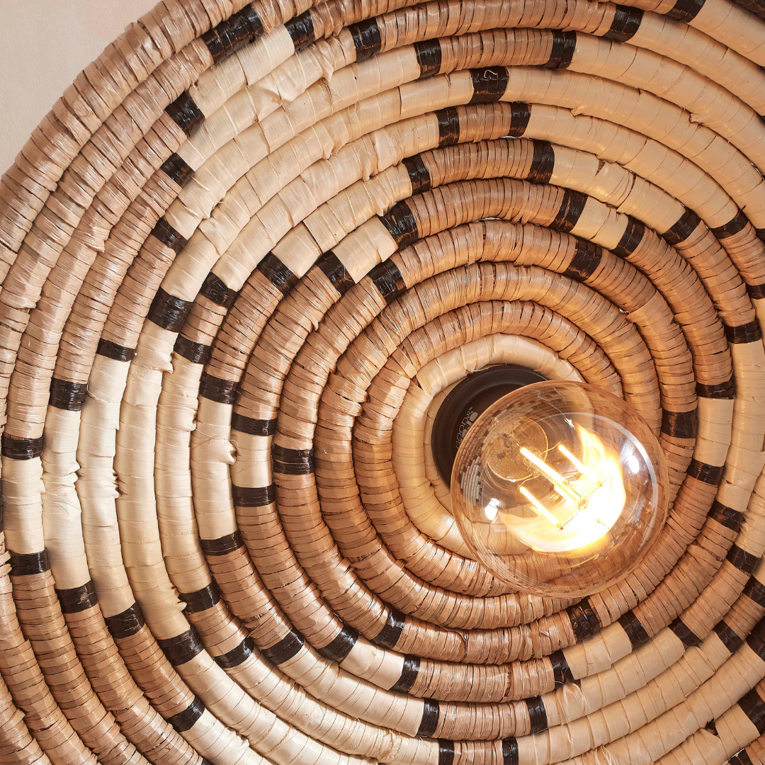 Ein Bild aus dem Taiga Store von taiga.green. Die Wandlampe Andros Diam.45x8cm verfügt über eine leuchtende, freiliegende Glühbirne in der Mitte eines kreisförmigen, geflochtenen Korbschirms mit Naturfasern und einem Spiralmuster in Hellbraun und Dunkelbraun. Jetzt bestellen im Eco Shop von www.taiga.green. Hier nachhaltig einkaufen mit dem Taiga Eco Rating