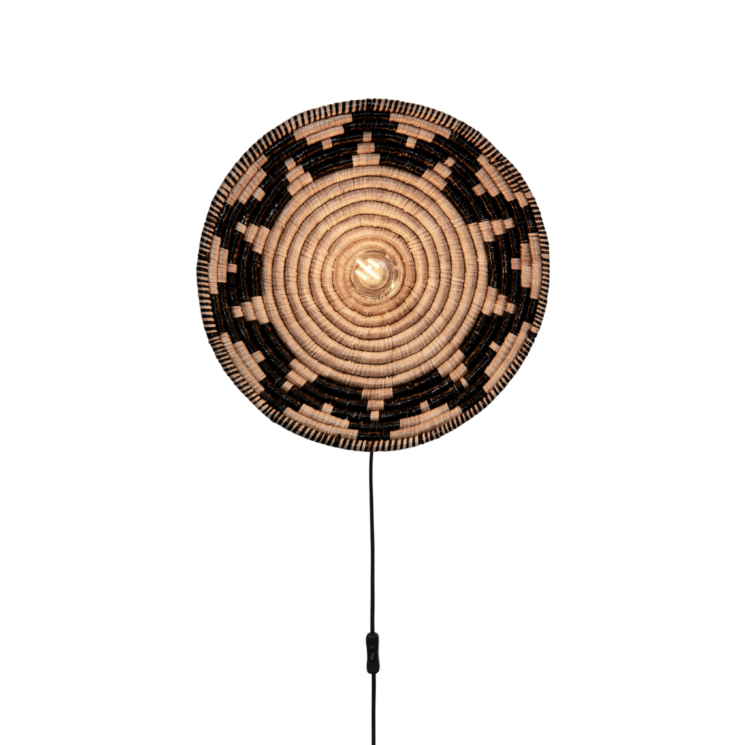 Ein Bild aus dem Taiga Store von taiga.green. Die Wandlampe Andros Diam.45x8cm ist eine runde Wandleuchte mit einer zentralen Glühbirne, die konzentrische Ringe und ein geometrisches schwarz-beiges Webmuster auf schwarzem Hintergrund aufweist. Das Stromkabel ist sichtbar aufgehängt. Jetzt bestellen im Eco Shop von www.taiga.green. Hier nachhaltig einkaufen mit dem Taiga Eco Rating