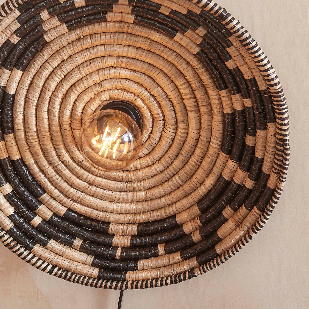 Ein Bild aus dem Taiga Store von taiga.green. Die Wandlampe Andros Diam.45x8cm ist eine runde Wandleuchte aus geflochtenem Rattan mit konzentrischen Kreisen in Natur und Schwarz, einer freiliegenden zentralen Glühbirne und wird an einer hellen Wand angebracht. Jetzt bestellen im Eco Shop von www.taiga.green. Hier nachhaltig einkaufen mit dem Taiga Eco Rating