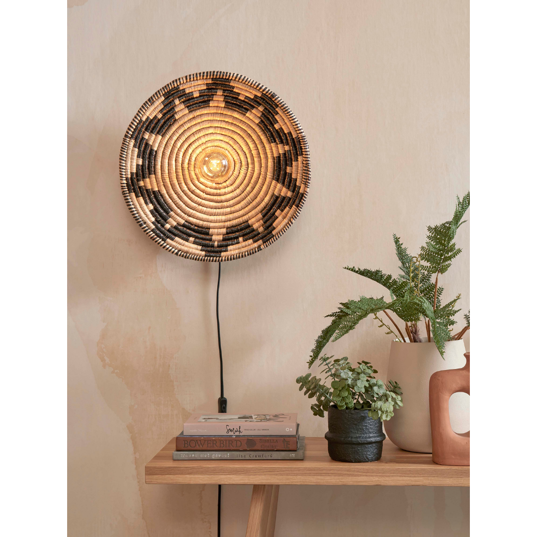 Ein Bild aus dem Taiga Store von taiga.green. Die Wandlampe Andros Diam.45x8cm mit spiralförmigem Flechtmuster leuchtet über einem Holzregal, das mit gestapelten Büchern, zwei kleinen Topfpflanzen und einer dekorativen Terrakotta-Vase mit Henkel dekoriert ist. Jetzt bestellen im Eco Shop von www.taiga.green. Hier nachhaltig einkaufen mit dem Taiga Eco Rating