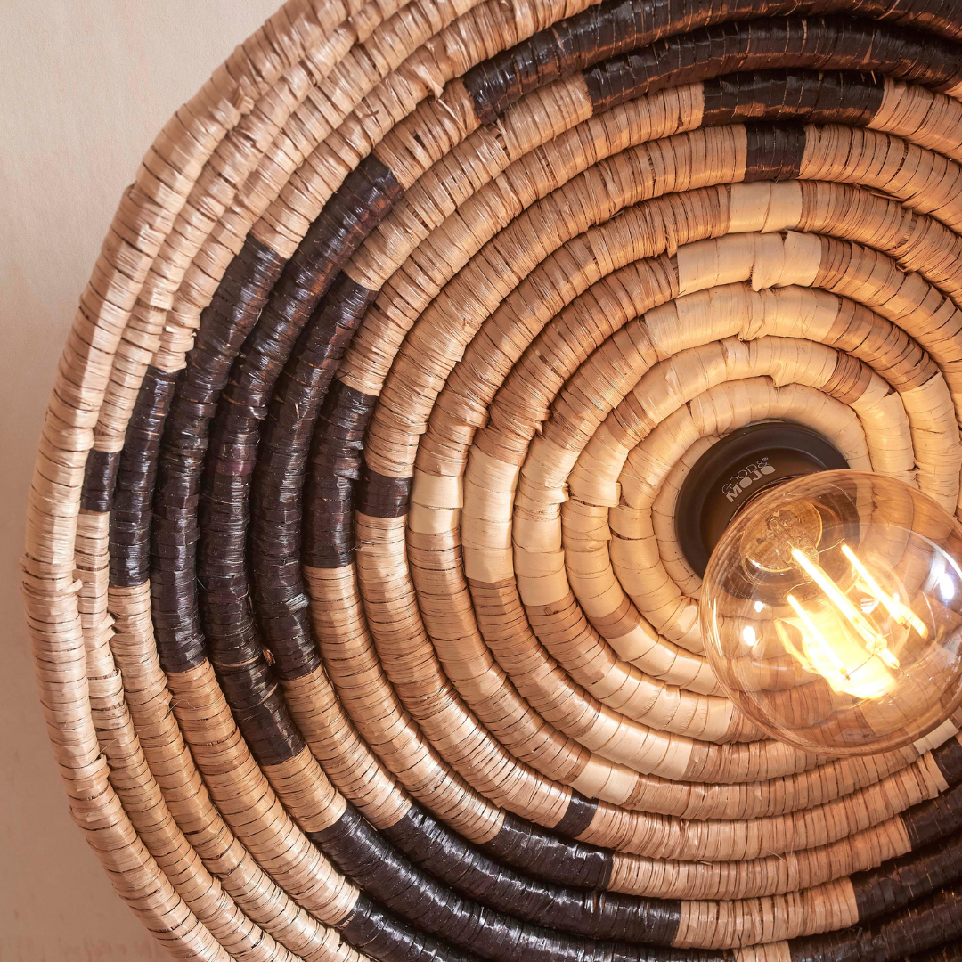 Ein Bild aus dem Taiga Store von taiga.green. Eine Nahaufnahme der Wandlampe Andros Diam.45x8cm, ein runder geflochtener Lampenschirm mit abwechselnd hellen und dunklen Streifen und einer leuchtenden Glühbirne in der Mitte rechts. Jetzt bestellen im Eco Shop von www.taiga.green. Hier nachhaltig einkaufen mit dem Taiga Eco Rating