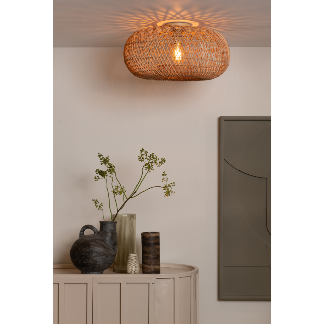 Ein Bild aus dem Taiga Store von taiga.green. Die Deckenlampe Arizona rattan/Schirm ø50x23cm S wirft gemusterte Schatten über ein helles Sideboard mit Vasen und einem Ast; rechts davon hängt ein abstraktes Wandkunstwerk. Jetzt bestellen im Eco Shop von www.taiga.green. Hier nachhaltig einkaufen mit dem Taiga Eco Rating