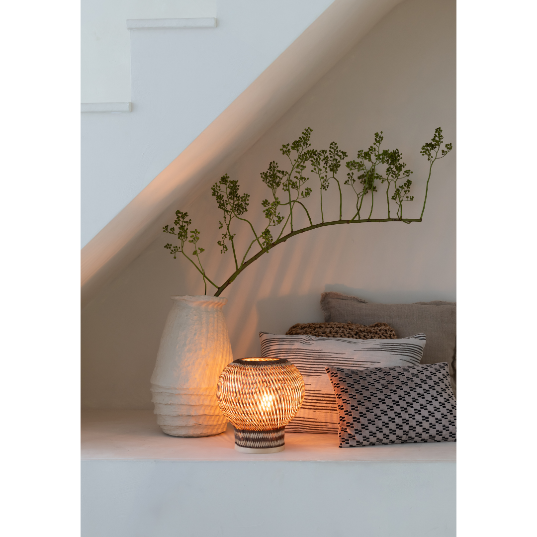 Ein Bild aus dem Taiga Store von taiga.green. Eine gemütliche Ecke unter der Treppe mit gestapelten, gemusterten Kissen, einer hohen, strukturierten weißen Vase mit einem grünen Zweig und der Tischlampe Barbados Bambus/Globe ø25xh.27cm, die warmes, gewobenes Licht ausstrahlt. Jetzt bestellen im Eco Shop von www.taiga.green. Hier nachhaltig einkaufen mit dem Taiga Eco Rating