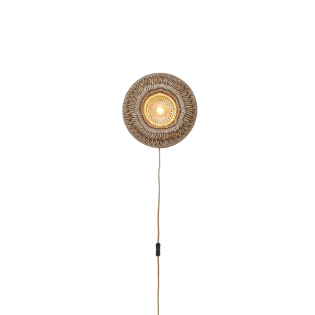 Ein Bild aus dem Taiga Store von taiga.green. Eine runde, geflochtene Wandlampe mit einer leuchtenden Glühbirne in der Mitte und einer Schnur, die gerade nach unten hängt, vor einem schlichten schwarzen Hintergrund. Jetzt bestellen im Eco Shop von www.taiga.green. Hier nachhaltig einkaufen mit dem Taiga Eco Rating