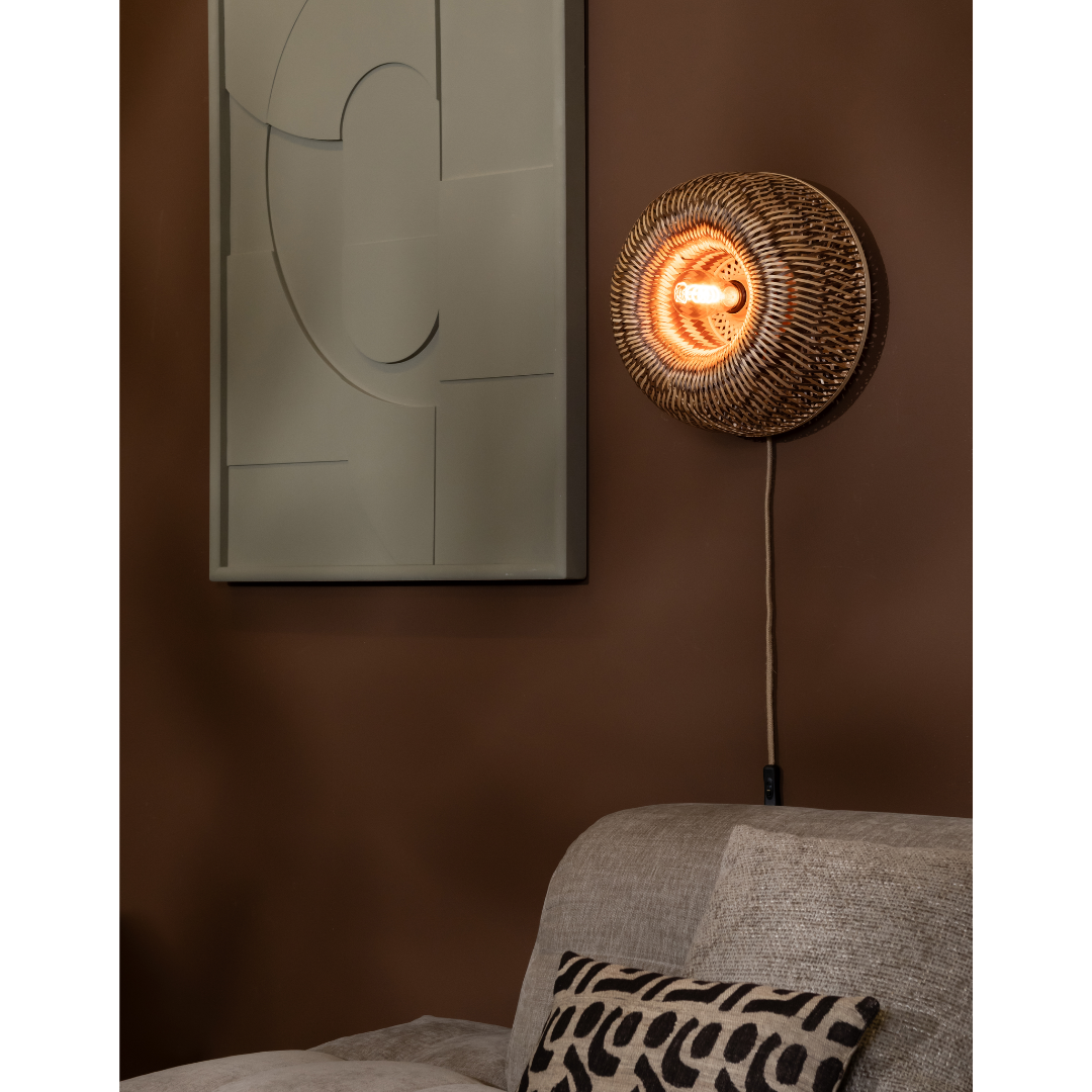Ein Bild aus dem Taiga Store von taiga.green. Eine runde Wandlampe aus Weidengeflecht leuchtet warm über einem beigen Sofa mit gemustertem Kissen; abstrakte Kunst in gedeckten Tönen hängt an einer braunen Wand in dem gemütlichen Wohnraum. Jetzt bestellen im Eco Shop von www.taiga.green. Hier nachhaltig einkaufen mit dem Taiga Eco Rating
