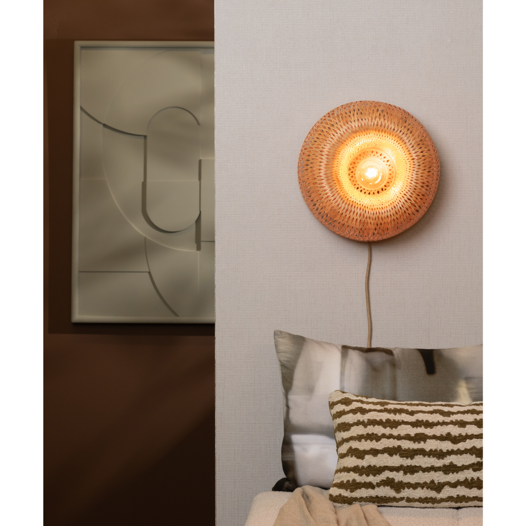 Ein Bild aus dem Taiga Store von taiga.green. Eine Wandlampe aus geflochtenem Rattan leuchtet warm über einem Bett mit gemusterten Kissen; ein modernes abstraktes Kunstwerk hängt an der braunen Wand neben dem Bett. Jetzt bestellen im Eco Shop von www.taiga.green. Hier nachhaltig einkaufen mit dem Taiga Eco Rating