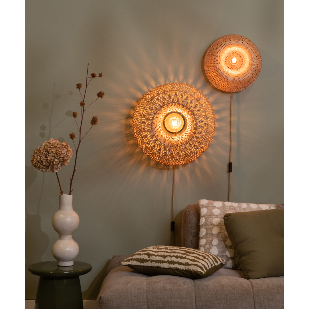 Ein Bild aus dem Taiga Store von taiga.green. Eine gemütliche Wohnzimmerszene mit zwei geflochtenen Wandlampen, die warme Lichtmuster werfen, einem beigen Sofa mit strukturierten Kissen und einem grünen Beistelltisch mit einer weißen Vase mit Trockenblumen. Jetzt bestellen im Eco Shop von www.taiga.green. Hier nachhaltig einkaufen mit dem Taiga Eco Rating