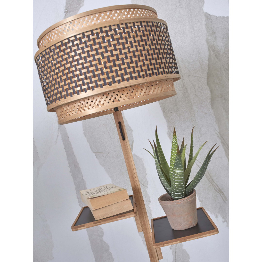 Ein Bild aus dem Taiga Store von taiga.green. Eine hölzerne Stehlampe mit einem geflochtenen, runden Lampenschirm steht neben einem kleinen schwarzen Regal mit einer Sukkulenten-Topfpflanze und zwei gestapelten Büchern vor einer hellen Wand mit abstrakten Mustern. Jetzt bestellen im Eco Shop von www.taiga.green. Hier nachhaltig einkaufen mit dem Taiga Eco Rating