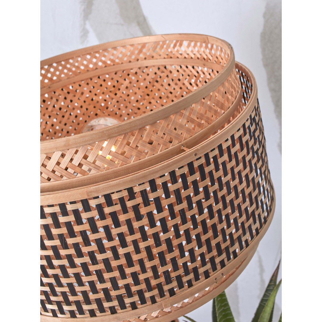 Ein Bild aus dem Taiga Store von taiga.green. Nahaufnahme eines Lampenschirms aus geflochtenem Rattan mit kreisförmigem, korbartigem Muster, das aus natürlichen und dunklen, verflochtenen Streifen besteht. Im Inneren ist eine Glühbirne zu sehen, und unten ist ein Teil einer Pflanze zu erkennen. Jetzt bestellen im Eco Shop von www.taiga.green. Hier nachhaltig einkaufen mit dem Taiga Eco Rating