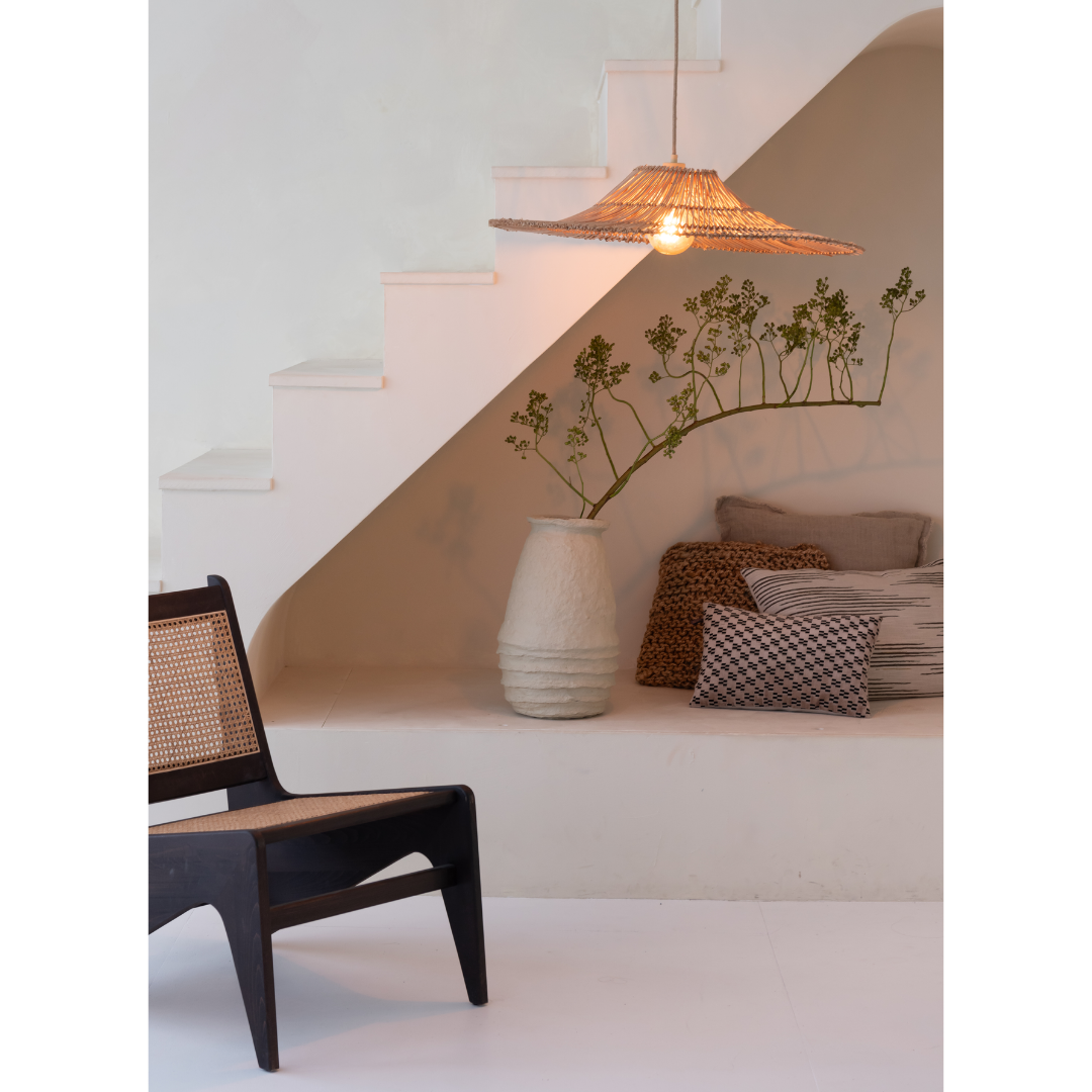 Ein Bild aus dem Taiga Store von taiga.green. Minimalistisches Interieur mit weißer Treppe, einer geflochtenen Hängeleuchte, einem schwarzen Stuhl mit Rohrrücken, einer großen Keramikvase mit Zweigen und verschiedenen neutralen Kissen, die in einer gemütlichen Ecke unter der Treppe angeordnet sind. Jetzt bestellen im Eco Shop von www.taiga.green. Hier nachhaltig einkaufen mit dem Taiga Eco Rating