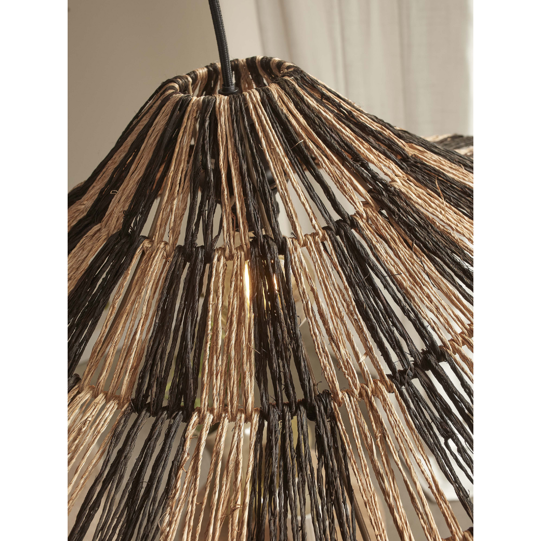 Ein Bild aus dem Taiga Store von taiga.green. Nahaufnahme eines gewebten Lampenschirms aus natürlichen und dunklen Fasern, die ein geometrisches, offenes Muster bilden, das das Licht durchscheinen lässt. Oben ist eine Kordel angebracht, und im unscharfen Hintergrund sind transparente Vorhänge zu sehen. Jetzt bestellen im Eco Shop von www.taiga.green. Hier nachhaltig einkaufen mit dem Taiga Eco Rating