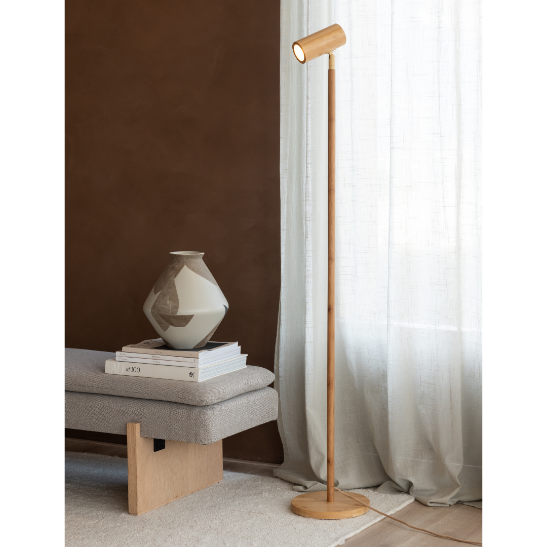 Ein Bild aus dem Taiga Store von taiga.green. Ein modernes, minimalistisches Zimmer mit einer hölzernen Stehlampe, einer grauen gepolsterten Bank, gestapelten Büchern und einer dekorativen Vase neben einem durchsichtigen weißen Vorhang und einer braunen Wand. Jetzt bestellen im Eco Shop von www.taiga.green. Hier nachhaltig einkaufen mit dem Taiga Eco Rating