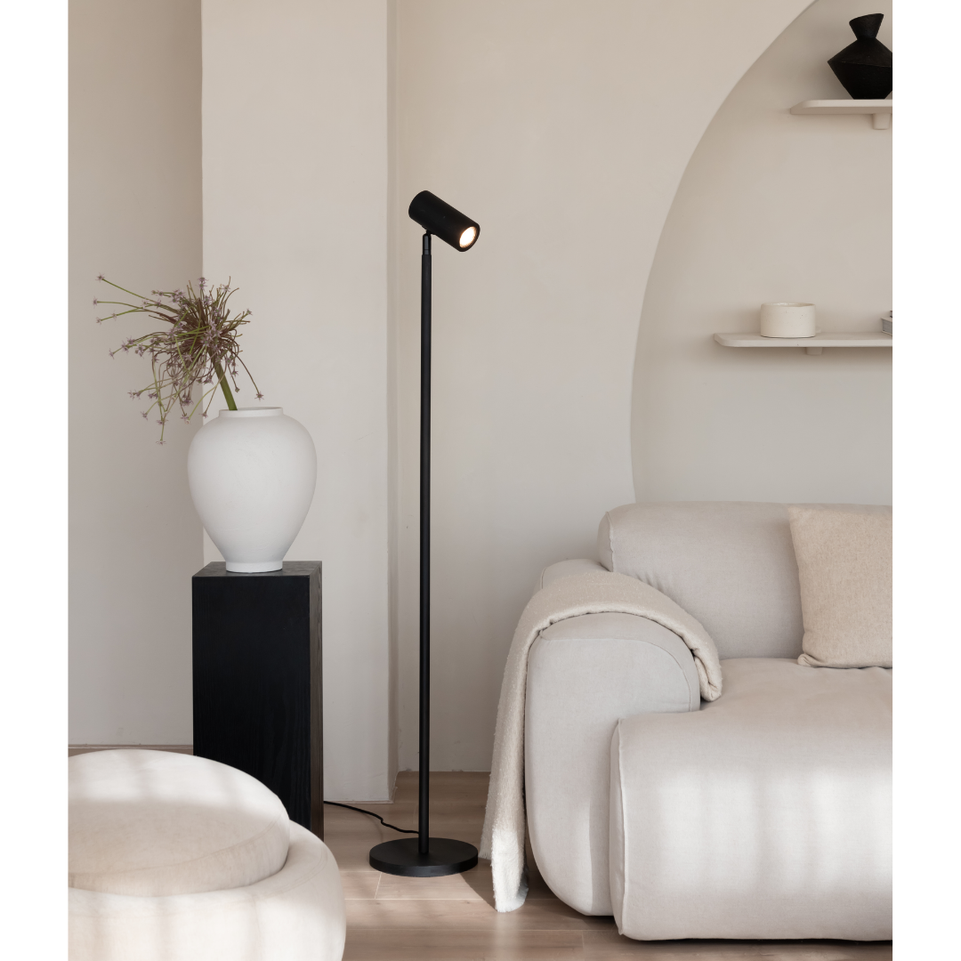 Ein Bild aus dem Taiga Store von taiga.green. Ein modernes Wohnzimmer mit einem hellbeigen Sofa, einer runden Ottomane, einer schwarzen Stehlampe und einer weißen Vase mit getrockneten Zweigen auf einem schwarzen Sockel. Minimalistisches Dekor und sanftes natürliches Licht erfüllen den Raum. Jetzt bestellen im Eco Shop von www.taiga.green. Hier nachhaltig einkaufen mit dem Taiga Eco Rating