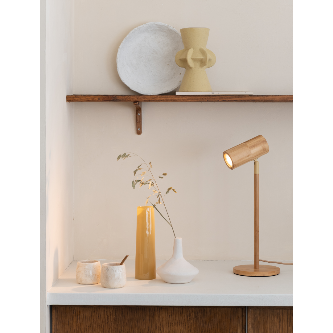 Ein Bild aus dem Taiga Store von taiga.green. Ein minimalistisch eingerichteter Schreibtisch mit einer Holzlampe, weißen und gelben Vasen, zwei kleinen Keramikgefäßen und getrockneten Pflanzenstängeln, an einer Wand mit einem Holzregal, auf dem ein weißer Teller und eine geometrische gelbe Skulptur stehen. Jetzt bestellen im Eco Shop von www.taiga.green. Hier nachhaltig einkaufen mit dem Taiga Eco Rating