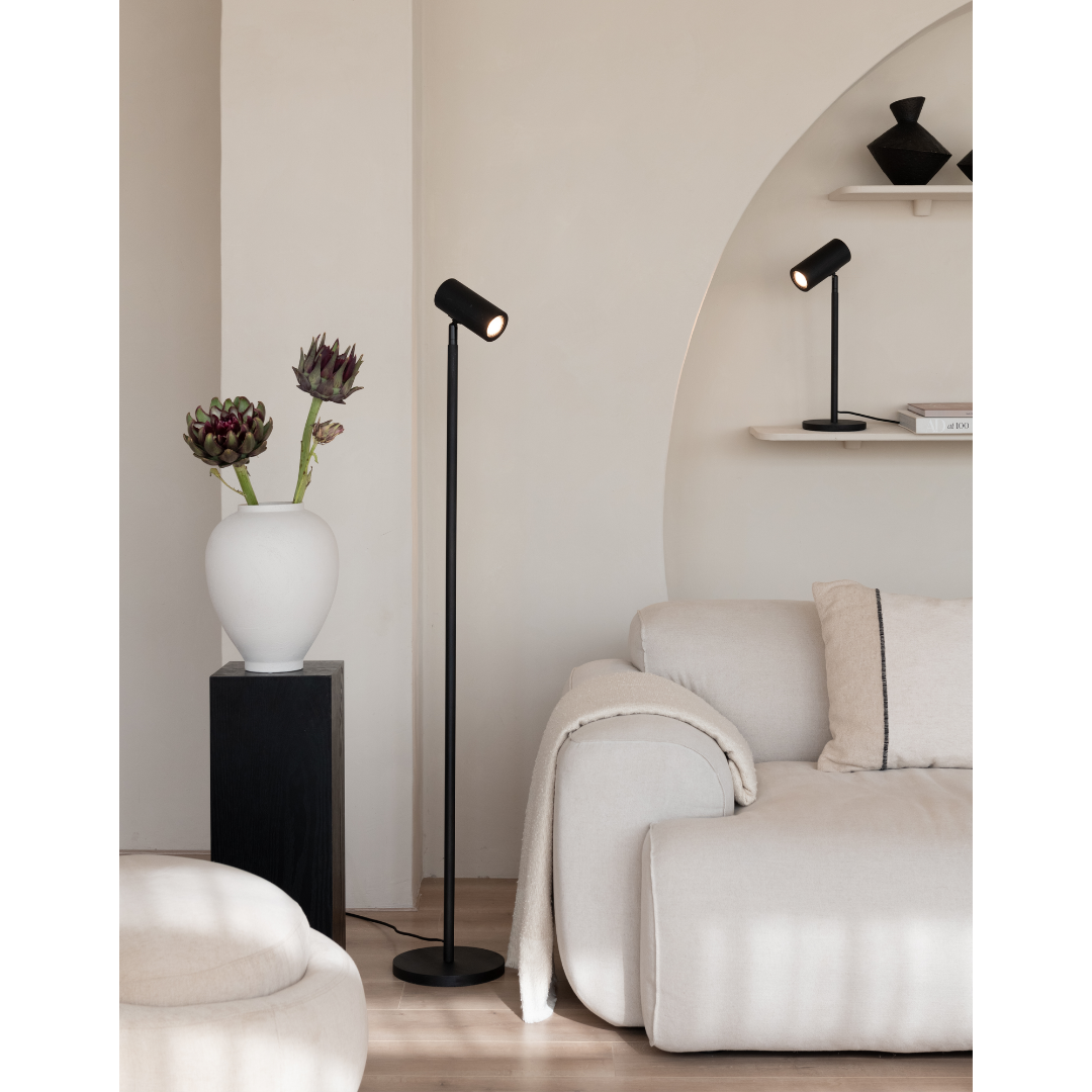 Ein Bild aus dem Taiga Store von taiga.green. Ein modernes Wohnzimmer mit einem beigen Sofa, einer schwarzen Stehlampe, einer weißen Vase mit Blumen auf einem schwarzen Sockel und einem Wandregal mit einer schwarzen Tischlampe und Dekorationsobjekten. Neutrale Farben und minimalistisches Dekor. Jetzt bestellen im Eco Shop von www.taiga.green. Hier nachhaltig einkaufen mit dem Taiga Eco Rating