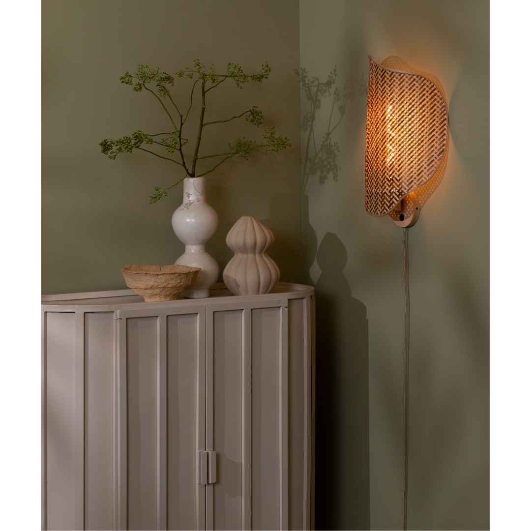 Ein Bild aus dem Taiga Store von taiga.green. Neben einem cremefarbenen Schrank mit einer weißen Vase mit grünen Zweigen, einer beigen Schale und einem skulpturalen Dekorationsstück leuchtet eine sanft beleuchtete geflochtene Wandlampe, alles vor einer gedämpften grünen Wand. Jetzt bestellen im Eco Shop von www.taiga.green. Hier nachhaltig einkaufen mit dem Taiga Eco Rating