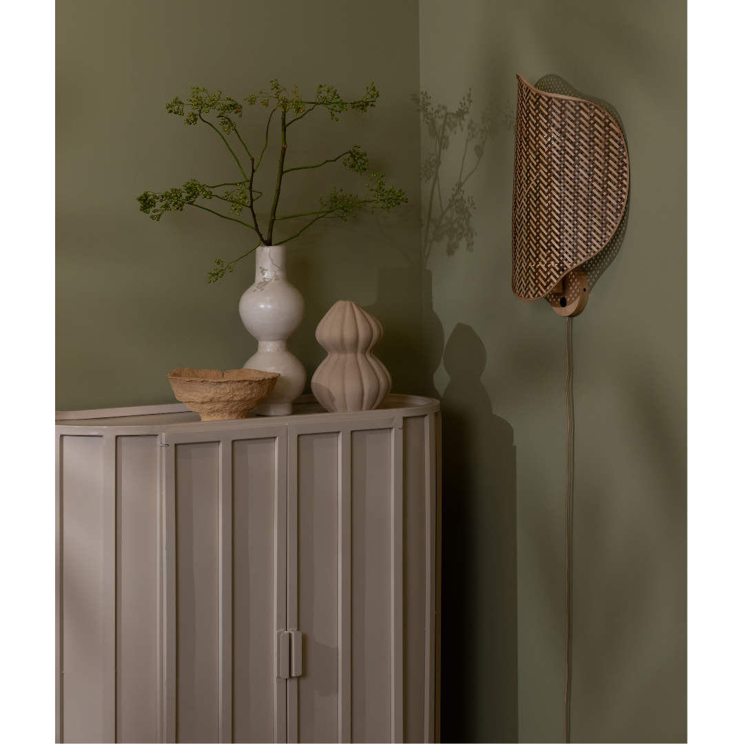 Ein Bild aus dem Taiga Store von taiga.green. Eine Ecke mit einem hellgrauen Schrank, einer weißen Vase mit grünen Zweigen, einer strukturierten Schale, einem beigen Dekorationsobjekt und einer geflochtenen Wandleuchte, die einen Schatten auf eine grüne Wand wirft. Jetzt bestellen im Eco Shop von www.taiga.green. Hier nachhaltig einkaufen mit dem Taiga Eco Rating