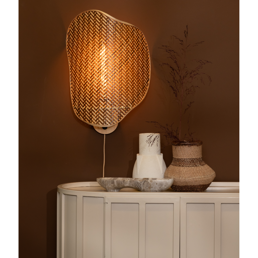 Ein Bild aus dem Taiga Store von taiga.green. Eine geflochtene Wandlampe spendet warmes Licht über einem weißen Schrank, in dem ein dekoratives Tablett mit gerollten Handtüchern und eine geflochtene Vase mit getrockneten Blättern stehen, alles vor einer braunen Wand. Jetzt bestellen im Eco Shop von www.taiga.green. Hier nachhaltig einkaufen mit dem Taiga Eco Rating