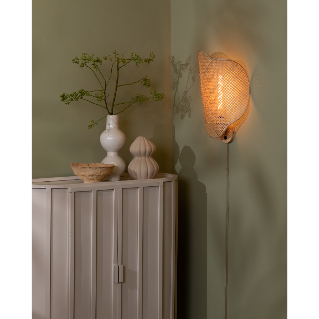 Ein Bild aus dem Taiga Store von taiga.green. Eine moderne Ecke mit einer hellgrünen Wand, einem weißen Schrank, einer weißen Vase mit Zweigen, einer geformten beigen Vase, einer kleinen geflochtenen Schale und einem dekorativen Wandleuchter, der ein warmes Licht ausstrahlt. Jetzt bestellen im Eco Shop von www.taiga.green. Hier nachhaltig einkaufen mit dem Taiga Eco Rating