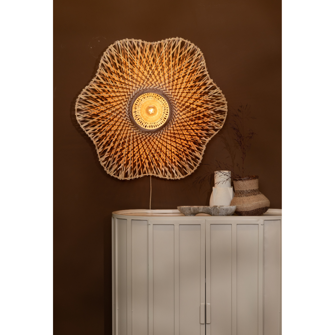 Ein Bild aus dem Taiga Store von taiga.green. Die Wandlampe Madura Bambus ø87x20cm L, eine große blumenförmige Rattanlampe, leuchtet warm über einem hellgrauen Schrank mit Vasen und getrockneten Pflanzen, der vor einer braunen Wand steht. Jetzt bestellen im Eco Shop von www.taiga.green. Hier nachhaltig einkaufen mit dem Taiga Eco Rating