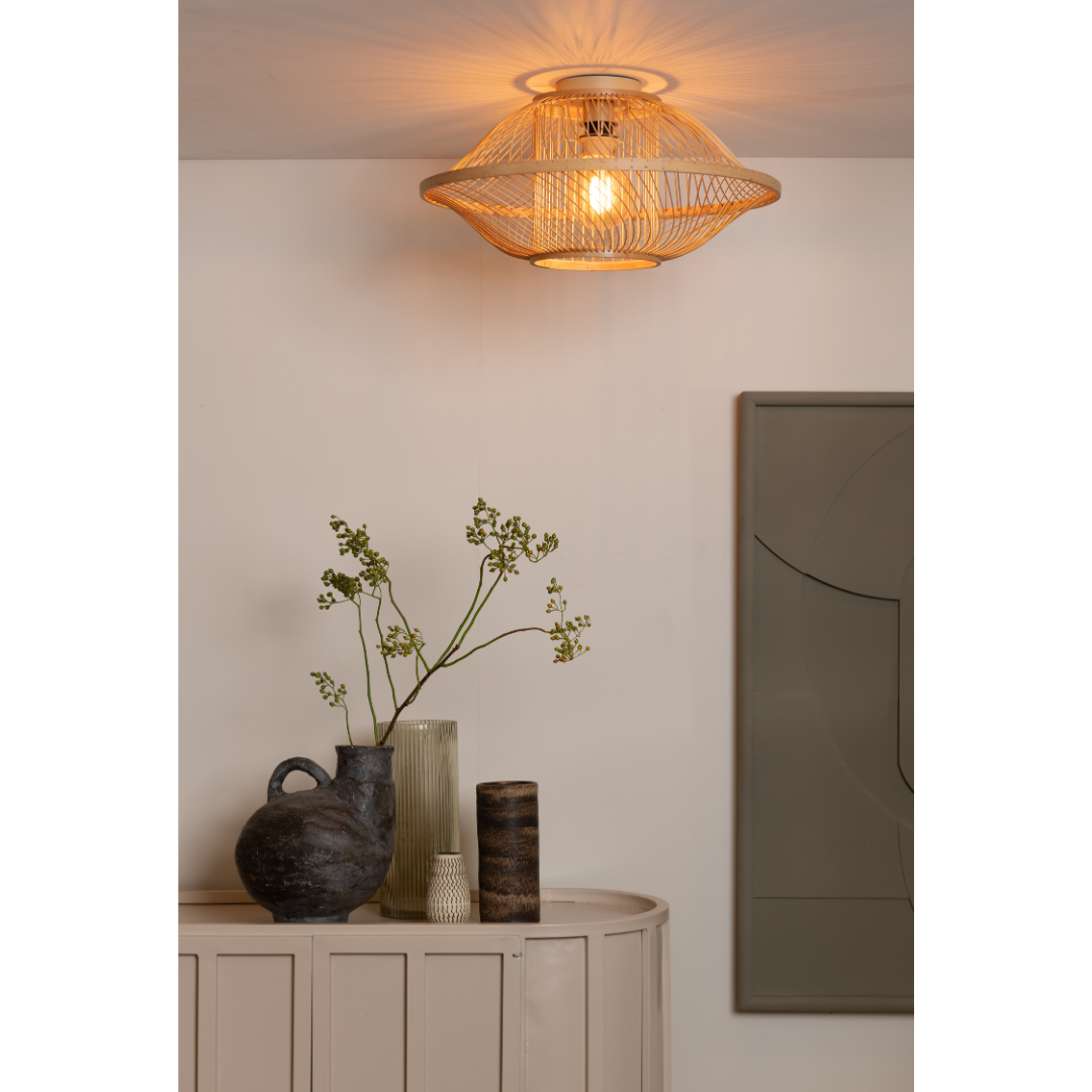 Ein Bild aus dem Taiga Store von taiga.green. Die Deckenlampe Maui Bambus/Schirm S, eine warme Deckenleuchte mit einem Schirm aus geflochtenem Rattan, leuchtet über einem beigen Sideboard, das mit Keramikvasen mit Grünpflanzen und strukturierten Mustern geschmückt ist. In der Nähe hängt ein großes abstraktes Wandgemälde. Jetzt bestellen im Eco Shop von www.taiga.green. Hier nachhaltig einkaufen mit dem Taiga Eco Rating