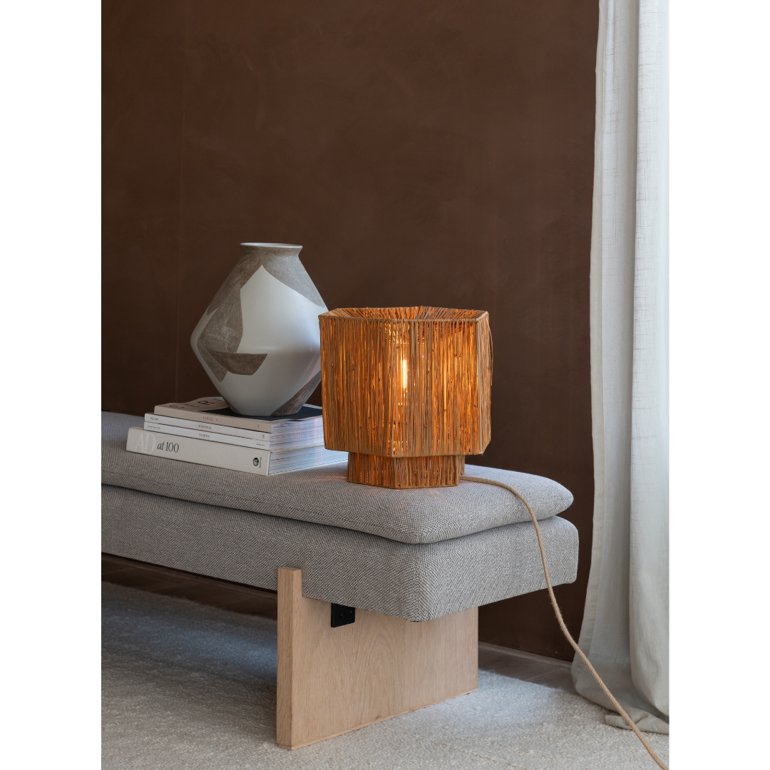 Ein Bild aus dem Taiga Store von taiga.green. Eine kleine Rattan-Tischlampe steht auf einer gepolsterten grauen Bank neben einer Keramikvase und gestapelten Büchern in einem gemütlichen Raum mit braunen Wänden und einem weißen Vorhang. Jetzt bestellen im Eco Shop von www.taiga.green. Hier nachhaltig einkaufen mit dem Taiga Eco Rating