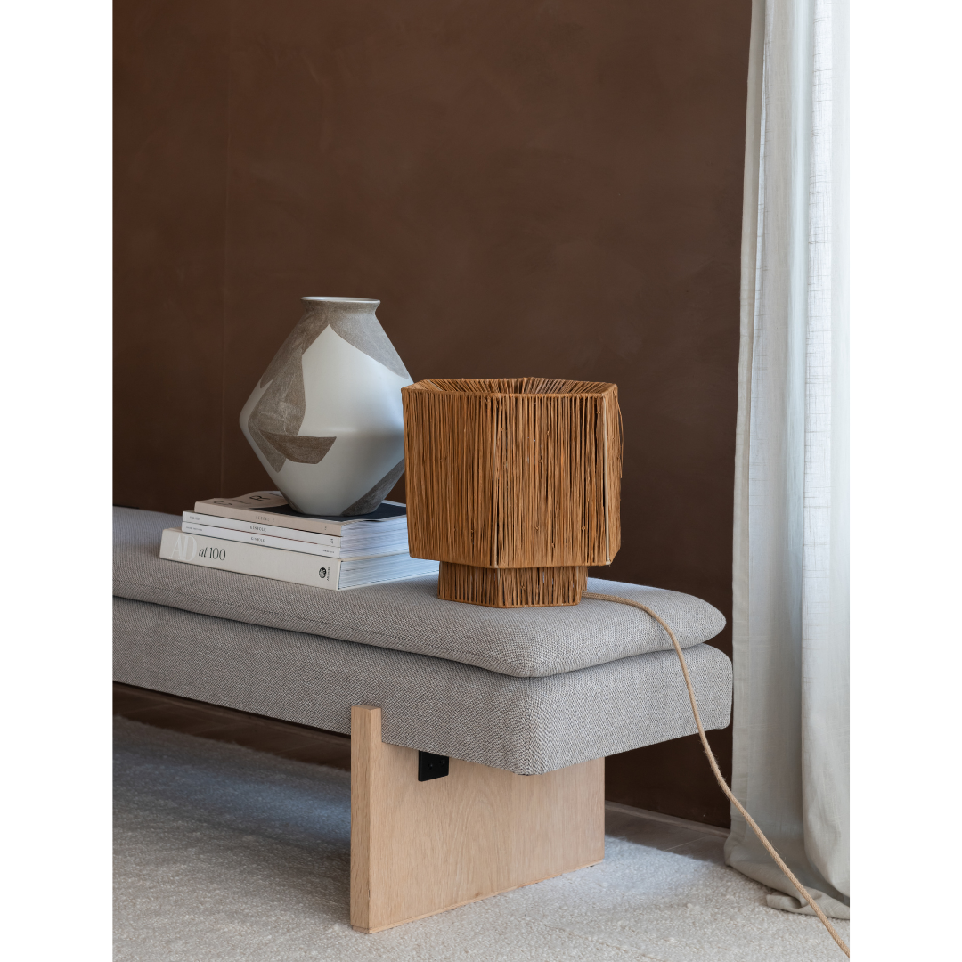 Ein Bild aus dem Taiga Store von taiga.green. Eine Tischlampe aus geflochtenem Rattan und eine Keramikvase stehen auf gestapelten Büchern auf einer hellgrauen Bank vor einer braunen Wand mit einem durchsichtigen weißen Vorhang daneben. Jetzt bestellen im Eco Shop von www.taiga.green. Hier nachhaltig einkaufen mit dem Taiga Eco Rating
