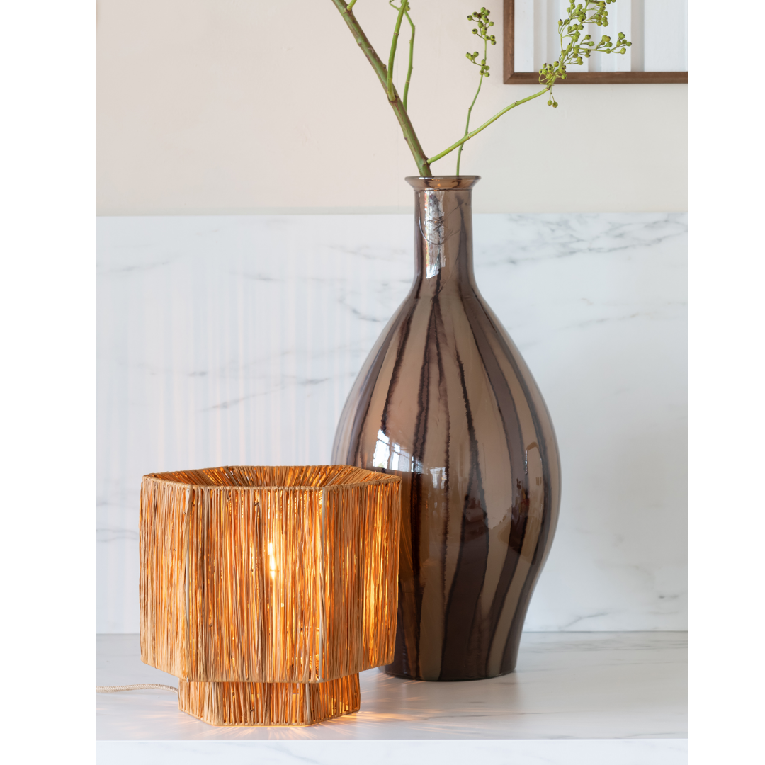 Ein Bild aus dem Taiga Store von taiga.green. Eine hohe, glänzende braune Vase mit dünnen vertikalen Streifen hält grüne Zweige neben einer leuchtenden, quadratischen Weidenlampe auf einer weißen Marmorfläche. Jetzt bestellen im Eco Shop von www.taiga.green. Hier nachhaltig einkaufen mit dem Taiga Eco Rating