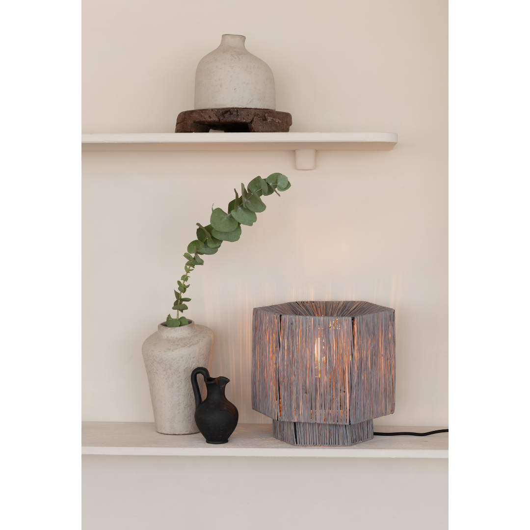Ein Bild aus dem Taiga Store von taiga.green. Auf einem dekorativen Regal stehen eine weiße Vase mit einem blättrigen Zweig, ein kleiner schwarzer Krug und eine geflochtene Lampe, die einen warmen Schein abgibt. Darüber befindet sich ein zweites Regal mit einem runden weißen Keramikobjekt auf einem Holzsockel. Jetzt bestellen im Eco Shop von www.taiga.green. Hier nachhaltig einkaufen mit dem Taiga Eco Rating