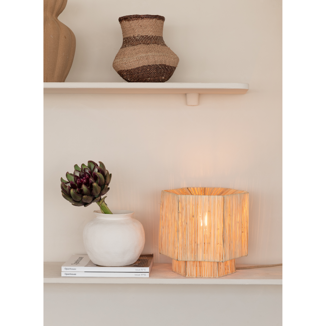 Ein Bild aus dem Taiga Store von taiga.green. Auf einem beigen Wandregal steht eine geflochtene Vase, während darunter eine weiße Keramikvase mit einer Artischocke neben einer leuchtenden Rattanlampe und zwei gestapelten Büchern auf einem unteren Regal steht. Jetzt bestellen im Eco Shop von www.taiga.green. Hier nachhaltig einkaufen mit dem Taiga Eco Rating