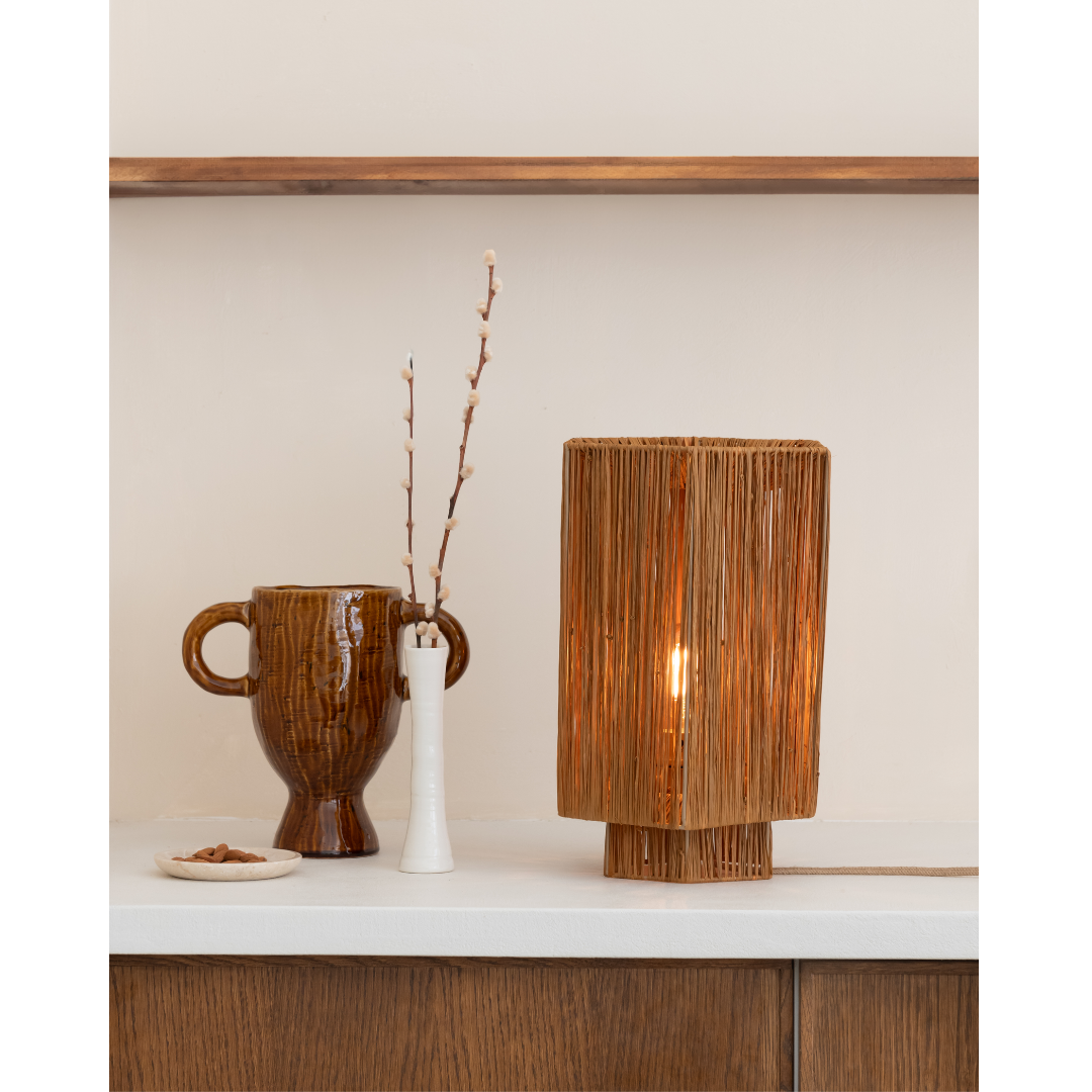 Ein Bild aus dem Taiga Store von taiga.green. Eine braune Keramikvase mit Henkeln, eine weiße Vase mit dünnen Zweigen und eine beleuchtete Korblampe stehen auf einer weißen Arbeitsplatte vor einer beigen Wand mit einem Holzregal darüber. Jetzt bestellen im Eco Shop von www.taiga.green. Hier nachhaltig einkaufen mit dem Taiga Eco Rating