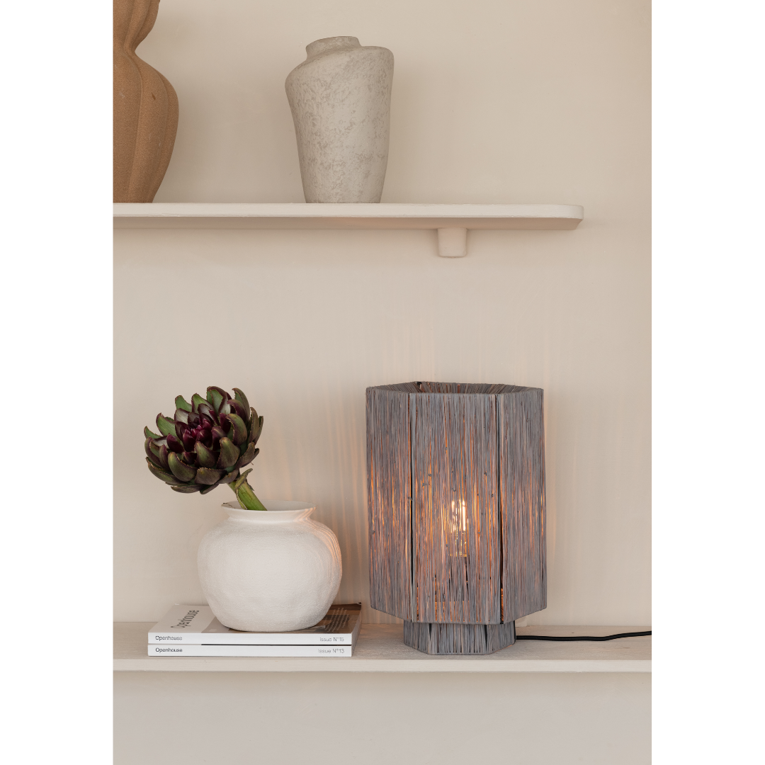 Ein Bild aus dem Taiga Store von taiga.green. Eine graue geflochtene Tischlampe steht auf einem Regal neben einer weißen Vase mit einer dunklen Artischocke, die auf zwei gestapelten Büchern steht. Eine weitere weiße Vase steht auf dem oberen Regal vor einer beigen Wand. Jetzt bestellen im Eco Shop von www.taiga.green. Hier nachhaltig einkaufen mit dem Taiga Eco Rating