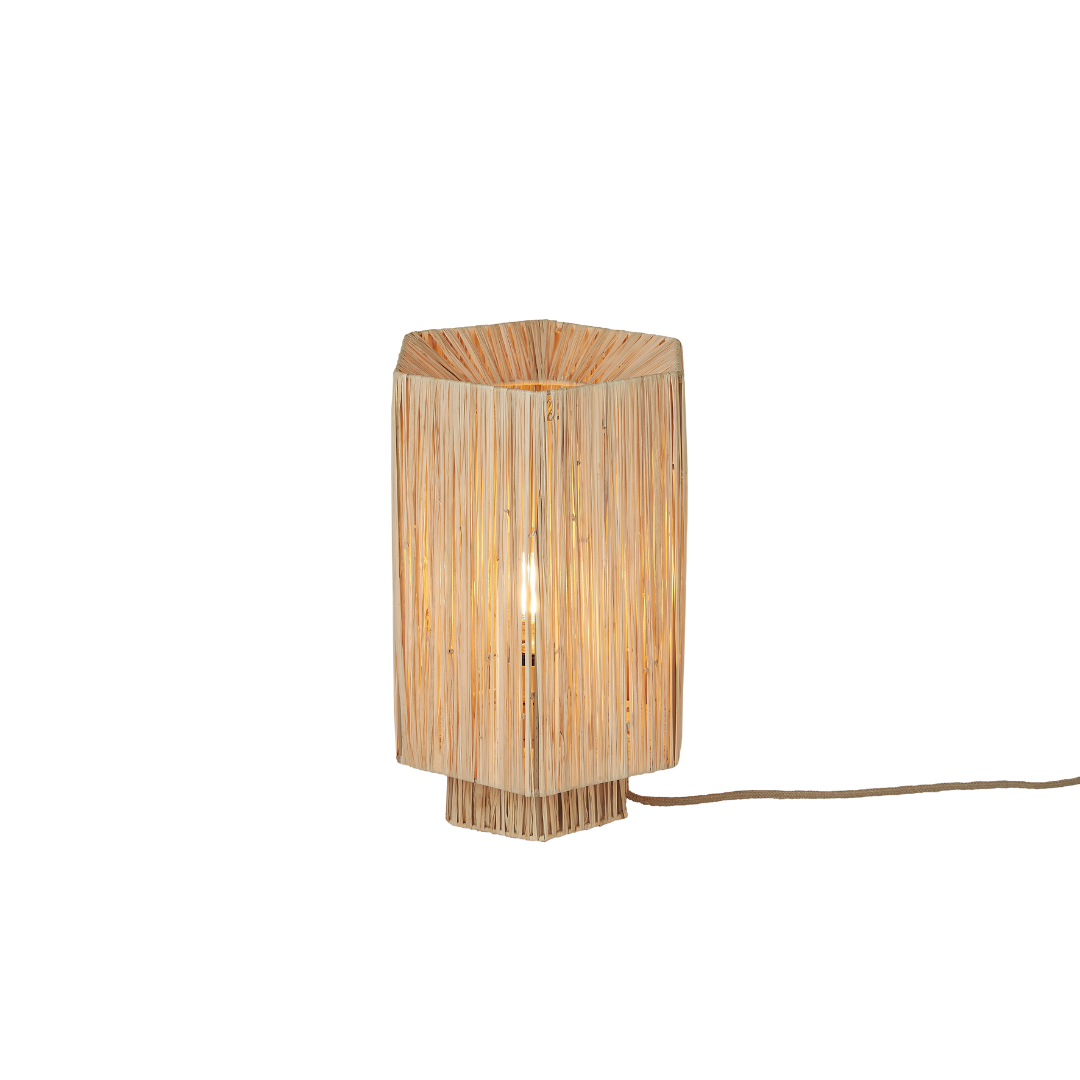 Ein Bild aus dem Taiga Store von taiga.green. Eine moderne Tischleuchte in sechseckiger Form mit einem Schirm aus gewebter Naturfaser, der ein warmes, weiches Licht abgibt. Die Lampe ist beleuchtet und hat ein sichtbares Kabel, das nach rechts verläuft. Jetzt bestellen im Eco Shop von www.taiga.green. Hier nachhaltig einkaufen mit dem Taiga Eco Rating