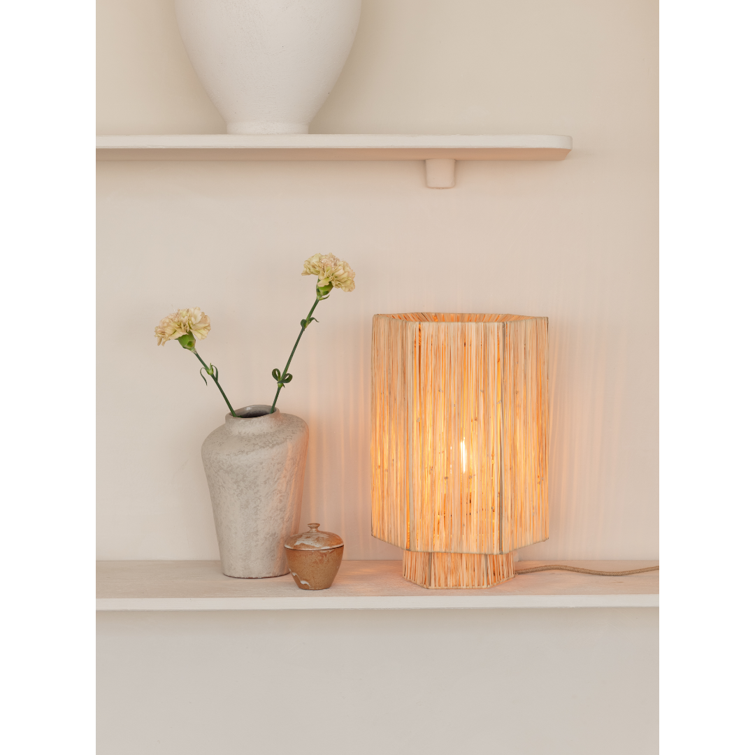 Ein Bild aus dem Taiga Store von taiga.green. Eine Keramikvase mit zwei gelben Blumen, ein kleines Glas mit Deckel und eine leuchtende Rattan-Tischlampe stehen auf einem hellen Regal vor einer beigen Wand. Jetzt bestellen im Eco Shop von www.taiga.green. Hier nachhaltig einkaufen mit dem Taiga Eco Rating