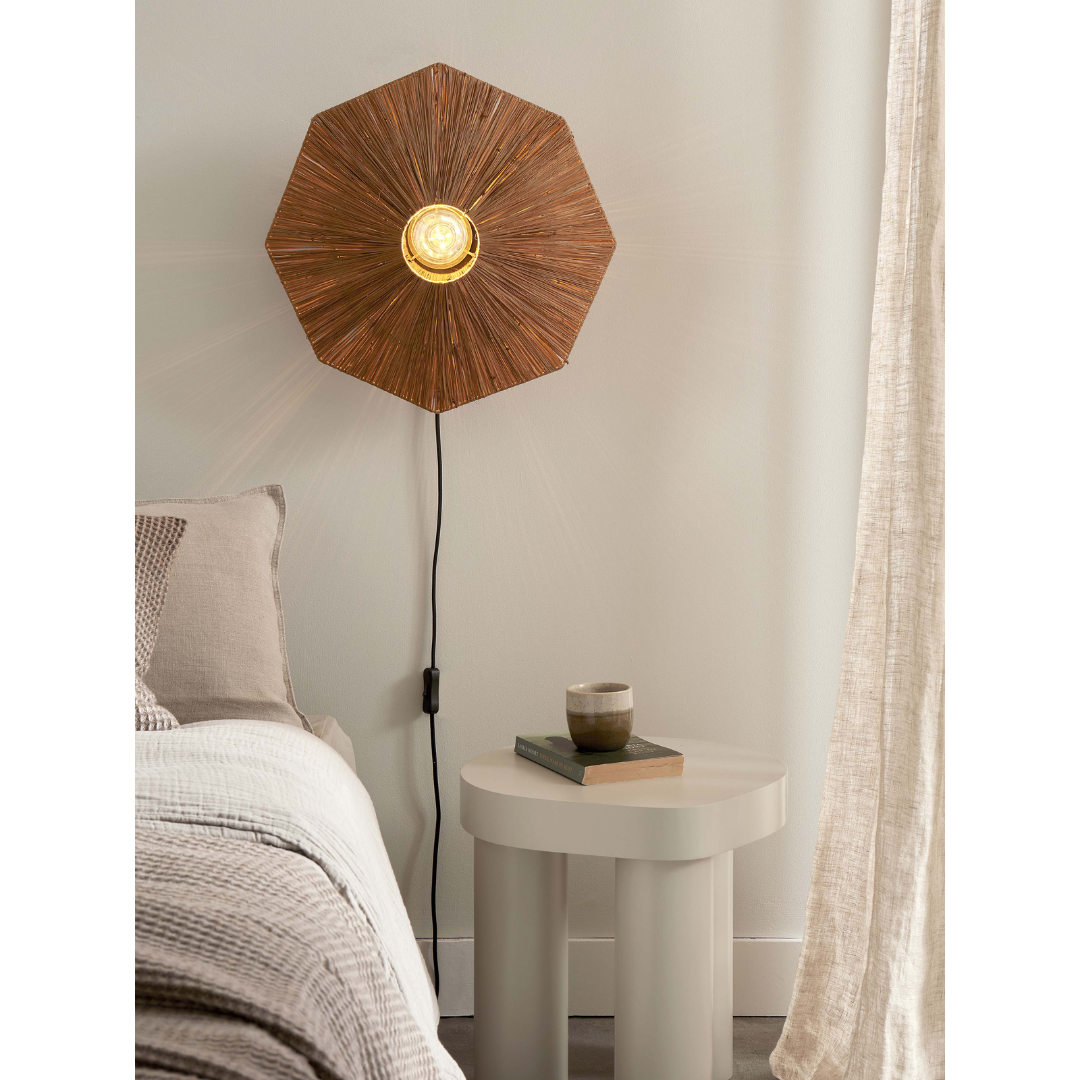 Ein Bild aus dem Taiga Store von taiga.green. Eine gemütliche Schlafzimmerszene mit einem Bett, einer strukturierten beigen Decke, einem runden weißen Nachttisch mit zwei Büchern und einer Tasse sowie einer an der Wand befestigten Sonnenbrillenlampe mit einem Holzschirm, die warm leuchtet. In der Nähe hängen hellbeige Vorhänge. Jetzt bestellen im Eco Shop von www.taiga.green. Hier nachhaltig einkaufen mit dem Taiga Eco Rating