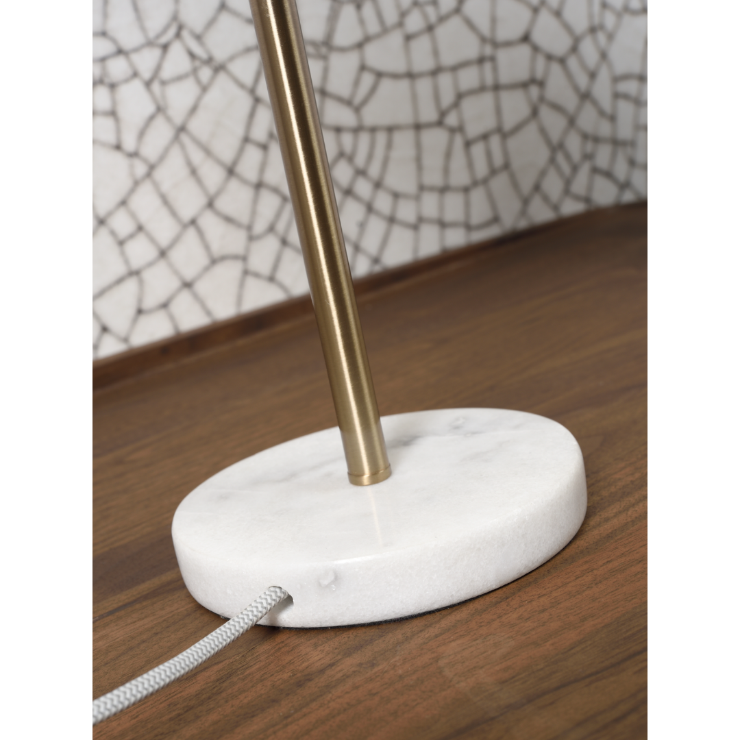 Ein Bild aus dem Taiga Store von taiga.green. Nahaufnahme einer Lampe mit einem runden weißen Marmorsockel und einem goldfarbenen Metallstiel, die auf einer hölzernen Fläche mit einer weißen Mosaikfliesenwand im Hintergrund steht. Vom Sockel geht eine mit Stoff überzogene Kordel aus. Jetzt bestellen im Eco Shop von www.taiga.green. Hier nachhaltig einkaufen mit dem Taiga Eco Rating