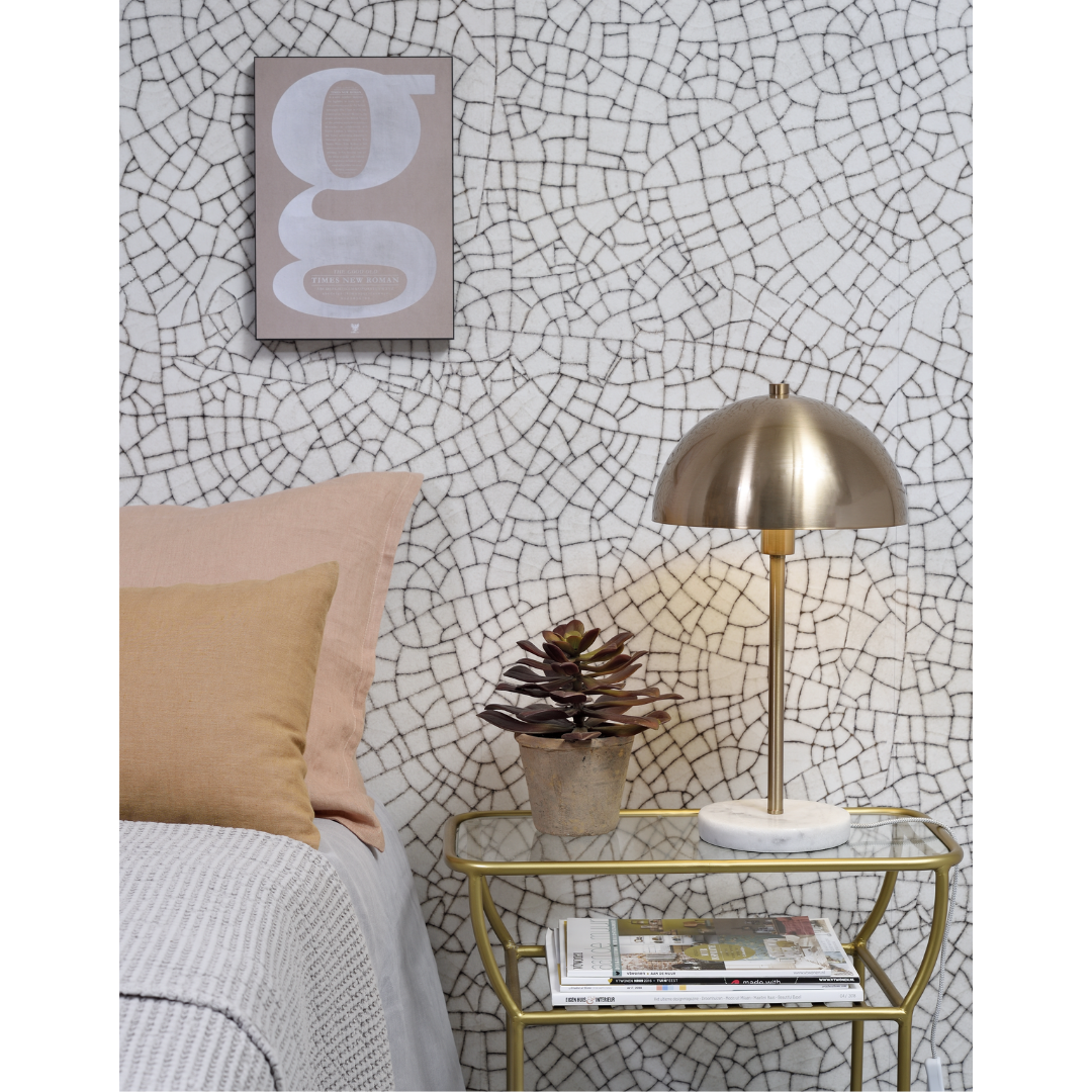 Ein Bild aus dem Taiga Store von taiga.green. Ein Nachttisch mit einer goldenen Lampe, einer kleinen Sukkulente und einem Stapel Zeitschriften steht neben einem Bett mit beigen Kissen. An der Wand hängt ein gerahmter Druck mit einem Mosaikfliesenmuster. Jetzt bestellen im Eco Shop von www.taiga.green. Hier nachhaltig einkaufen mit dem Taiga Eco Rating