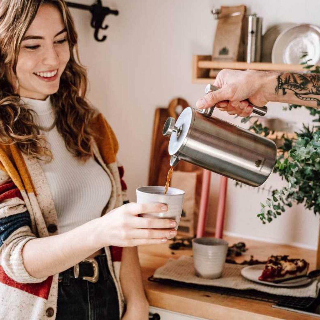 Taiga Store Eine Frau mit langen braunen Haaren lächelt als ihr eine Tasse Kaffee aus einer Metallkanne eingeschenkt wird Sie trägt einen gestreiften Pullover und steht in einer gemütlichen Küche mit Pflanzen und Gebäck auf der Theke bestellen bei wwwtaigagreen Shop