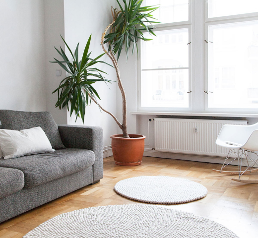 Ein Bild aus dem Taiga Store von taiga.green. Helles, minimalistisches Wohnzimmer mit grauem Sofa, weißem Kissen, großer Topfpflanze und weißem Schaukelstuhl. Zwei runde gewebte Teppiche auf Holzboden und große Fenster lassen Tageslicht herein. Jetzt bestellen im Eco Shop von www.taiga.green. Hier nachhaltig einkaufen mit dem Taiga Eco Rating