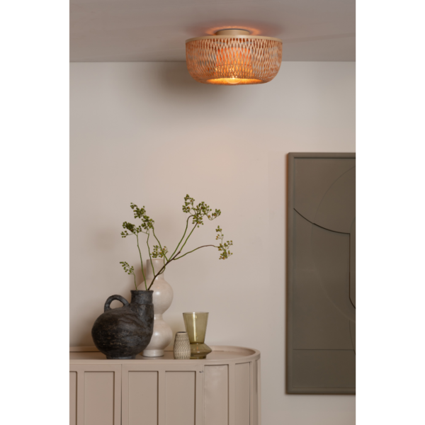 Ein Bild aus dem Taiga Store von taiga.green. Die Deckenlampe Barbados Bambus leuchtet warm über einem Sideboard mit Keramikvasen, einem grünen Glas und einem blattreichen Zweig. In der Nähe ziert ein neutrales abstraktes Kunstwerk die Wand. Jetzt bestellen im Eco Shop von www.taiga.green. Hier nachhaltig einkaufen mit dem Taiga Eco Rating