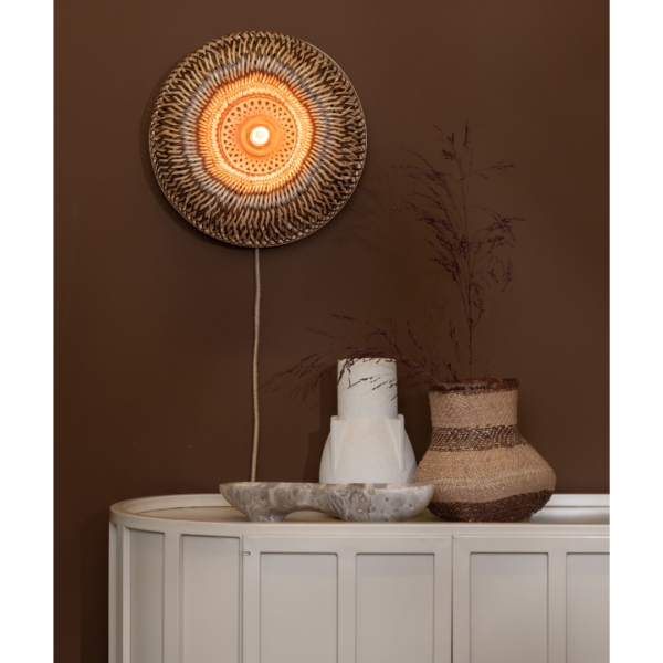 Ein Bild aus dem Taiga Store von taiga.green. Eine runde geflochtene Wandlampe leuchtet warm über einem weißen Konsolentisch mit dekorativen Vasen und einer Schale vor einer braunen Wand mit getrockneten Blättern in einer Vase. Jetzt bestellen im Eco Shop von www.taiga.green. Hier nachhaltig einkaufen mit dem Taiga Eco Rating