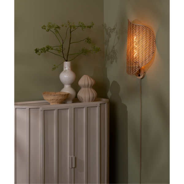 Ein Bild aus dem Taiga Store von taiga.green. Neben einem cremefarbenen Schrank mit einer weißen Vase mit grünen Zweigen, einer beigen Schale und einem skulpturalen Dekorationsstück leuchtet eine sanft beleuchtete geflochtene Wandlampe, alles vor einer gedämpften grünen Wand. Jetzt bestellen im Eco Shop von www.taiga.green. Hier nachhaltig einkaufen mit dem Taiga Eco Rating