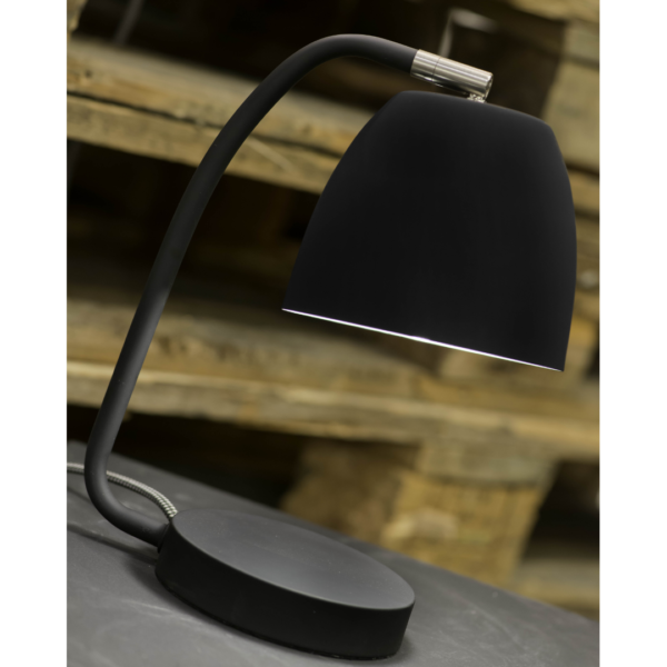 Ein Bild aus dem Taiga Store von taiga.green. Eine moderne schwarze Schreibtischlampe mit gebogenem Hals und kuppelförmigem Schirm, beleuchtet und auf eine dunkle Fläche gestellt. Im unscharfen Hintergrund sind Holzpaletten zu sehen. Jetzt bestellen im Eco Shop von www.taiga.green. Hier nachhaltig einkaufen mit dem Taiga Eco Rating
