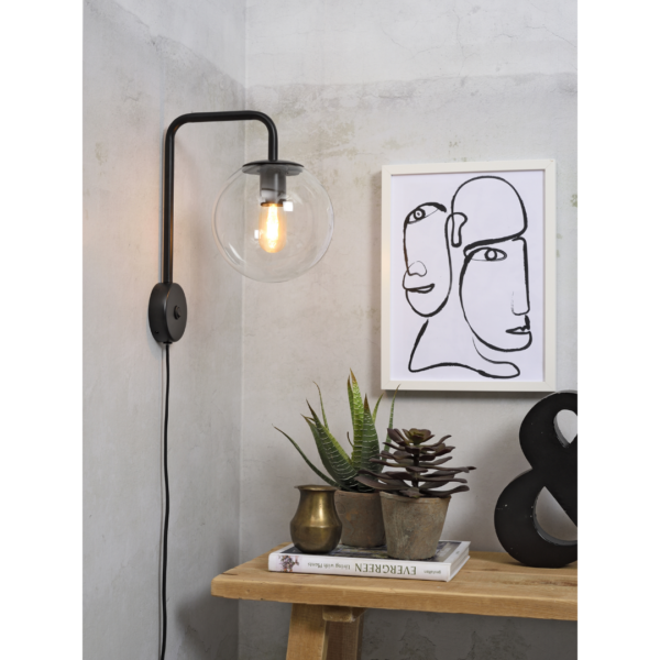 Ein Bild aus dem Taiga Store von taiga.green. Eine moderne Wandlampe mit rundem Glasschirm leuchtet über einem Holztisch mit Topfpflanzen, Messingvasen, einem schwarzen Und-Zeichen und einer gerahmten abstrakten Gesichtszeichnung an einer hellen Wand. Jetzt bestellen im Eco Shop von www.taiga.green. Hier nachhaltig einkaufen mit dem Taiga Eco Rating