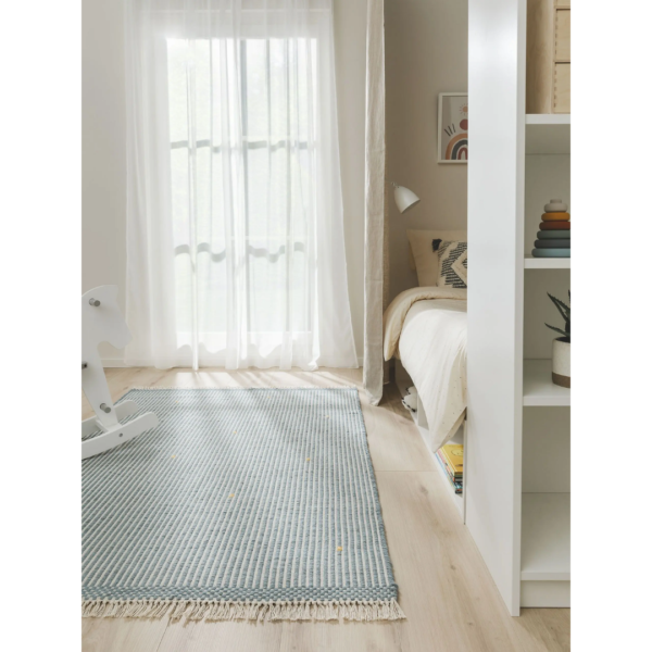 Ein Bild aus dem Taiga Store von taiga.green. Helles, gemütliches Schlafzimmer mit einem großen Fenster, das von transparenten Vorhängen verdeckt wird, einem blau gestreiften Teppich, einem kleinen Bett mit gemusterten Kissen, einem weißen Schaukelpferd und einem weißen Bücherregal mit Büchern und einer Topfpflanze. Jetzt bestellen im Eco Shop von www.taiga.green. Hier nachhaltig einkaufen mit dem Taiga Eco Rating
