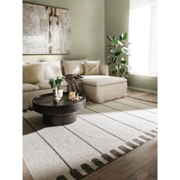Ein Bild aus dem Taiga Store von taiga.green. Ein gemütliches Wohnzimmer mit einem beigen Sektionssofa, weichen Kissen, einem runden schwarzen Couchtisch mit Kerzen, einer Topfpflanze, großer abstrakter Wandkunst und einem gemusterten Teppich in Naturtönen. Licht strömt durch ein Fenster herein. Jetzt bestellen im Eco Shop von www.taiga.green. Hier nachhaltig einkaufen mit dem Taiga Eco Rating