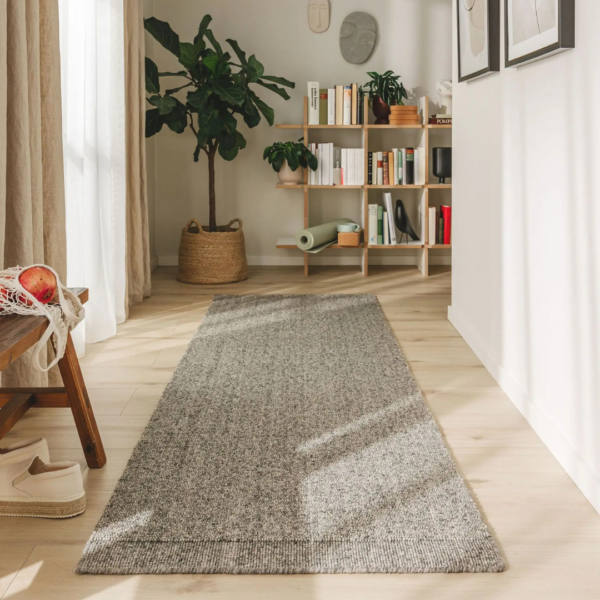 Ein Bild aus dem Taiga Store von taiga.green. Ein sonnendurchfluteter Flur mit hellem Holzboden, einem grauen Läufer, einer Topfpflanze, einer Holzbank mit einer Netztasche und Hausschuhen und einem Bücherregal mit Büchern und Dekoration an der Rückwand. Jetzt bestellen im Eco Shop von www.taiga.green. Hier nachhaltig einkaufen mit dem Taiga Eco Rating